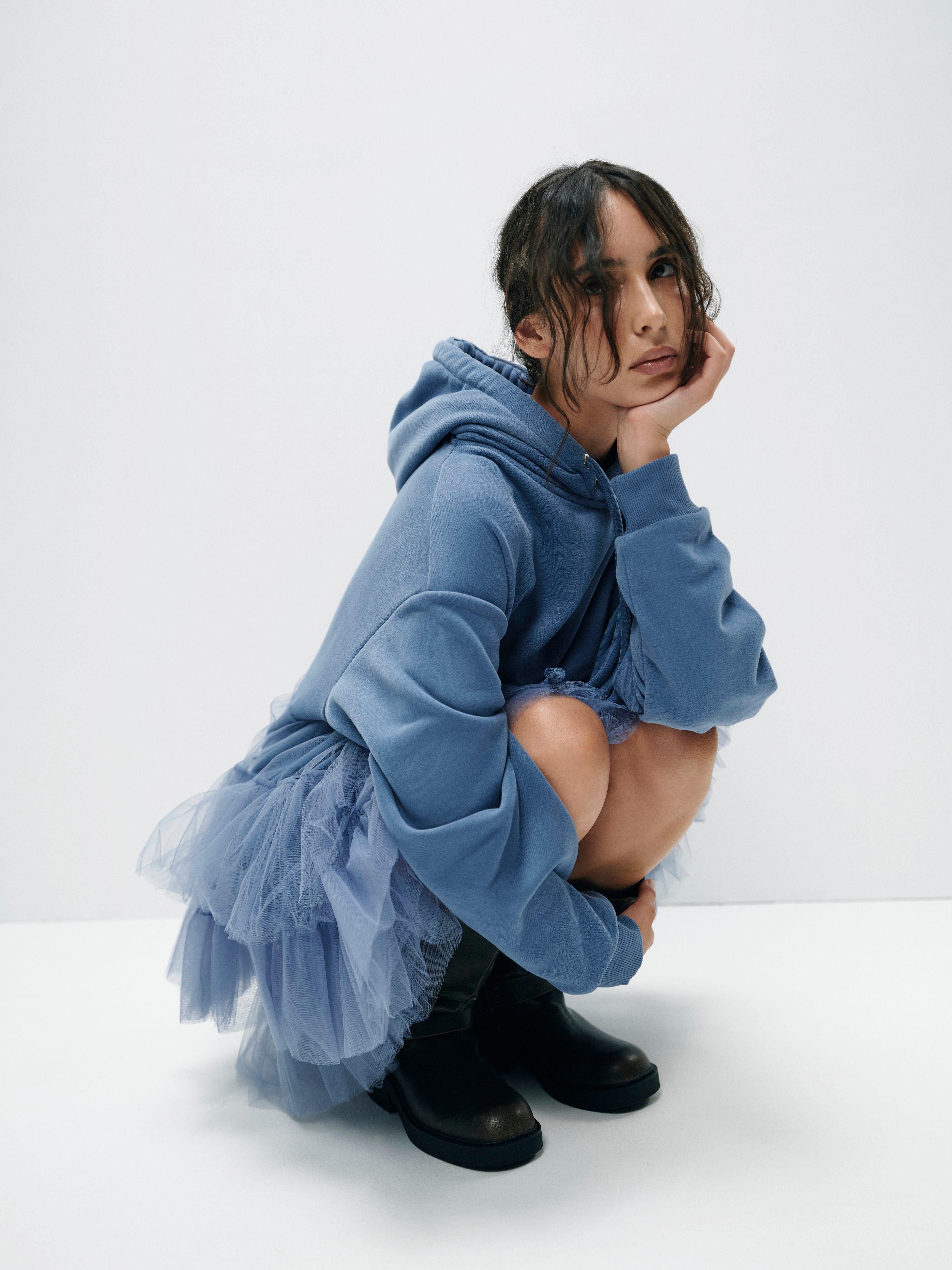 Model sitting in hoodie dress with dual drawstrings and layered tulle, demonstrating the relaxed yet stylish design with a voluminous, airy effect